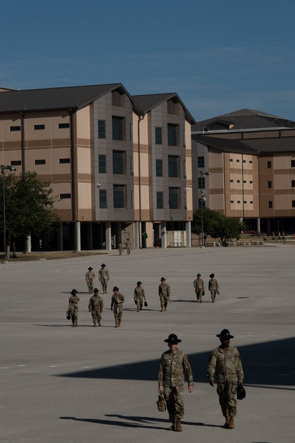 Military training instructors in campaign hats walk across a sunlit parade ground in front of barracks