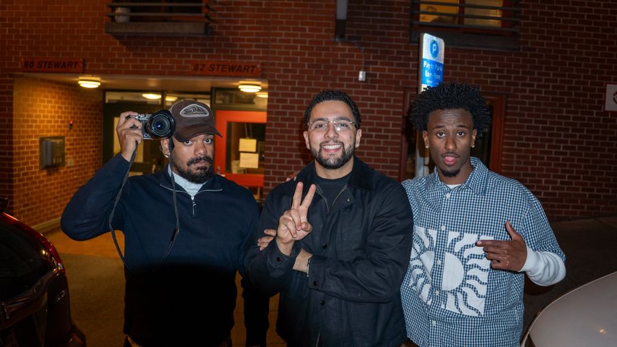 Three friends pose on a city street at night under warm light, one holding a camera up